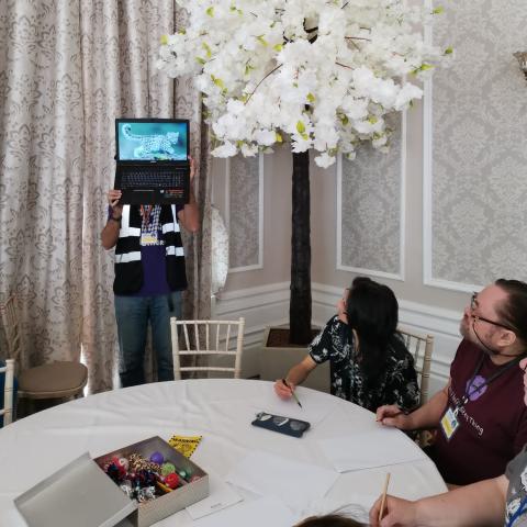 A person holds up a laptop for others to see what to copy – blindfolded!